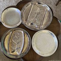 Four silver-plated serving trays displayed on a wooden table, two oval sectional trays with branch-like dividers, one smaller embossed round tray, one larger plain round tray.