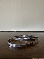 Three stacked sterling silver bracelets on a wooden surface, visible distinct engraving patterns.