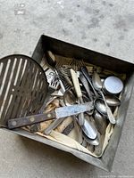Top view of mixed cutlery and kitchen utensils inside a metal container showing spoons, forks, knife, slotted spatula, and small metal container.