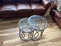 Two round metal and glass tables with overlapping design placed in front of leather sofas on wood flooring.