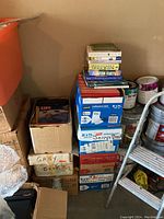 Boxes stacked in a garage corner with labeled boxes such as "Gary's Books" and visible book spines on top of the boxes, next to paint cans and a ladder.