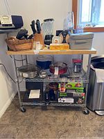 Front view of metal shelving unit with butcher block top loaded with kitchen items including knives, blender, decorative box, and plastic cups.
