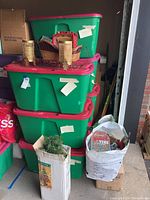 Five stacked green plastic storage bins with red lids, boxed artificial Christmas tree leaning against bins, holiday bags and boxes on the floor nearby. Items stored in a garage setting.