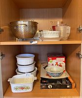 Top shelf with metal colander, glass pitcher, glass measuring cup, and CorningWare casserole dish.