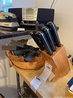 Five kitchen knives with black handles stored in a wooden block on a kitchen counter. Alongside, a woven basket contains several knife sharpening rods with black handles.
