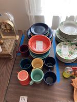 Photo showing stacked dinner plates, bread plates with green floral pattern, and colorful Fiestaware mugs on a table with a decorative clock nearby.