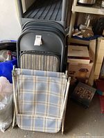 Four black metal folding chairs with padded striped seats stacked together with two folded lawn chaise lounges with light blue and beige checkered fabric, set inside a storage area.