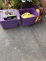 Two purple plastic totes with lids outdoors, containing hairstyling tools and accessories. Babyliss hair dryer box on one tote.