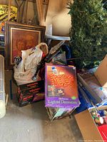Photo of assorted Halloween and holiday decorations in garage including lighted display box, garden inflatable box, artificial tree, plastic tote, and framed artwork.