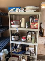 Overview photo of 4-tier plastic shelving unit filled with assorted kitchenware and small appliances, showing plates, bowls, pitchers, boxed vacuum cleaner, and various kitchen accessories and appliances.