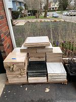 Stacked marble tiles in beige, black, and white outdoors. Roughly square shape and various sizes. Tiles appear heavy and stored on wooden pallets with some torn paper wrappings.