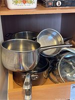 View of various sizes of stainless steel sauce pans stacked together along with glass lids and steamer insert inside a wooden shelf.