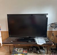 Front view of Sharp Aquos 52 inch LCD TV on wooden table with manual, remotes and other items visible below.
