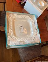 White square ceramic platter with raised lattice braided border inside a Tiffany blue box with tissue paper on a wooden chair and table.