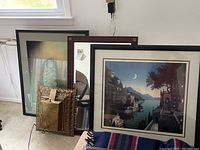 Four framed pieces arranged leaning against a wall including a signed art print with broken glass, framed light box, framed mirror, and coastal town wall art.