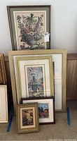 Multiple framed prints stacked up against a wall, showing various print and frame styles, mainly floral and architectural themes.
