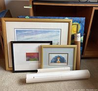 Five framed prints of varied sizes arranged against furniture and floor with three white rolled art tubes in front