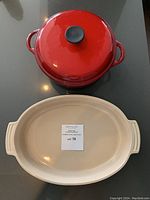 Top-down view of red cast iron pot with lid and green oval open casserole dish with chip, both on black surface, lot label visible