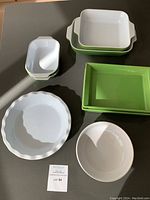 Full view of all cookware items including green casserole dishes, white pie plate, white bowl, and three small white rectangular dishes on a grey surface.