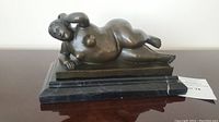 Side view of bronze reclining nude female figure on a two-tier black marble base showing overall form and base wear.
