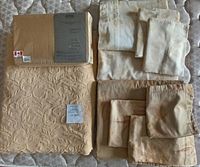 All bed linens displayed on mattress: folded quilt, sheets, duvet cover, shams, and throw cushion cover in various gold tones and patterns.