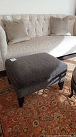 Side view of the charcoal grey upholstered ottoman placed in front of a couch with beige upholstery and two pillows, on a patterned rug.