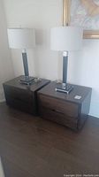 Two matching grey stained wood bedside tables side by side on hardwood floor with lamps on top