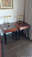 Two wooden side tables placed side-by-side with brass lamps on top, showing shape and finish.