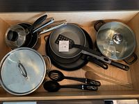 Full shot of all pots, pans, utensils in wooden drawer showing various sized pots with lids and frying pans with utensils.