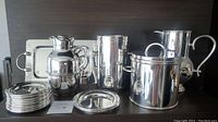 Full view of silver plated barware including water jug, ice bucket, wine cooler, coffee jug, tray, 10 coasters, and 2 bottle holders on dark shelf