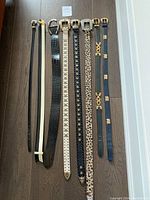 Full view of eight women's belts laid out on a wood floor showing various colors, buckles, and styles from thin to wide.