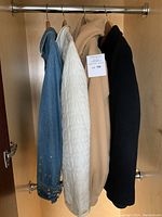 Photo showing four coats hanging in a closet sleeve, from left: blue denim jean jacket, white quilted jacket, camel peacoat, and black wool coat.