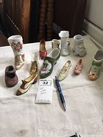 Photo of a collection of small ceramic and decorative shoe figurines in varied colors and styles arranged on a table with a pen and auction card for scale.