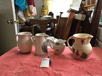 Four ceramic pitchers of different sizes and designs arranged on a pink cloth surface in a room.