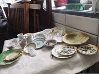 Photo of assorted single china pieces including cups, saucers, and plates with varied colors and floral designs, showing condition including chips and wear, arranged on table.