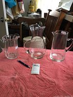 Three clear glass vessels on pink textured cloth: left scalloped rim container, center large round beer pitcher with handle, right tall clear pitcher with etched design