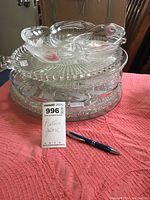 Stack of assorted clear glass platters and trays on pink fabric, showing various patterns and shapes including round and sectioned platters, plus a pen for scale.