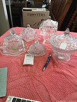 Full view of five various glass covered candy and butter dishes arranged on a textured pink cloth.