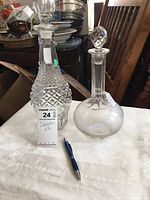 Two glass decanters placed side by side on a table with a pen for scale. One decanter is textured with a diamond pattern, the other is smooth with etched detail. Both have glass stoppers.