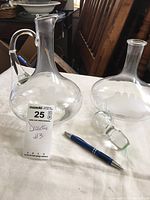 Two glass decanters shown side by side on a white surface, one with stopper and etched ship design, the other with etched grapes and leaves, with a pen for scale.