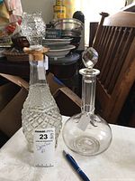 Pair of clear glass decanters with stoppers. One has a diamond cut pattern with a gold rim below stopper, the other has a rounded smooth base with vertical lined neck and starburst base detail.