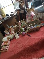 Photo showing a variety of Easter decorations including large bunny figurine in pink bonnet, smaller bunny decorations, decorative eggs, framed bunny picture and Easter-themed plate on a red tablecloth.