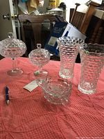 Full view of Fostoria Cubist glassware on a pink quilted table cover showing the lidded candy dishes, two vases, and candle holders.