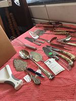 Photo showing an array of kitchen utensils including pie servers, spatulas, whisk, and ladle arranged on a red cloth.
