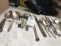 Assorted flatware including large serving spoons, regular spoons, forks, and one child fork from Campbell's, laid out on a patterned white tablecloth.