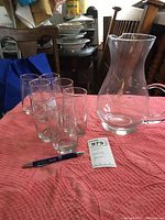 Clear glass pitcher with handle and six matching clear glass tumblers on a pink cloth.