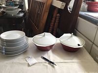 Stack of white plates with thin red rim next to two red and white covered casserole dishes on table with a pen for scale.