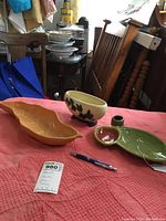Three Roseville pottery items displayed on a red quilt; orange leaf-shaped bowl, beige ivy planter, and green leaf-shaped tray with a round compartment.
