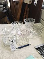 Eight clear glass small bowls arranged on a white fabric surface with wood chair and some other background items.