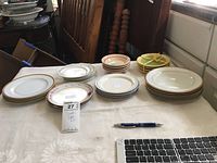 Multiple stacks of small plates of varying floral and plain designs on a table with labels and a pen for scale.